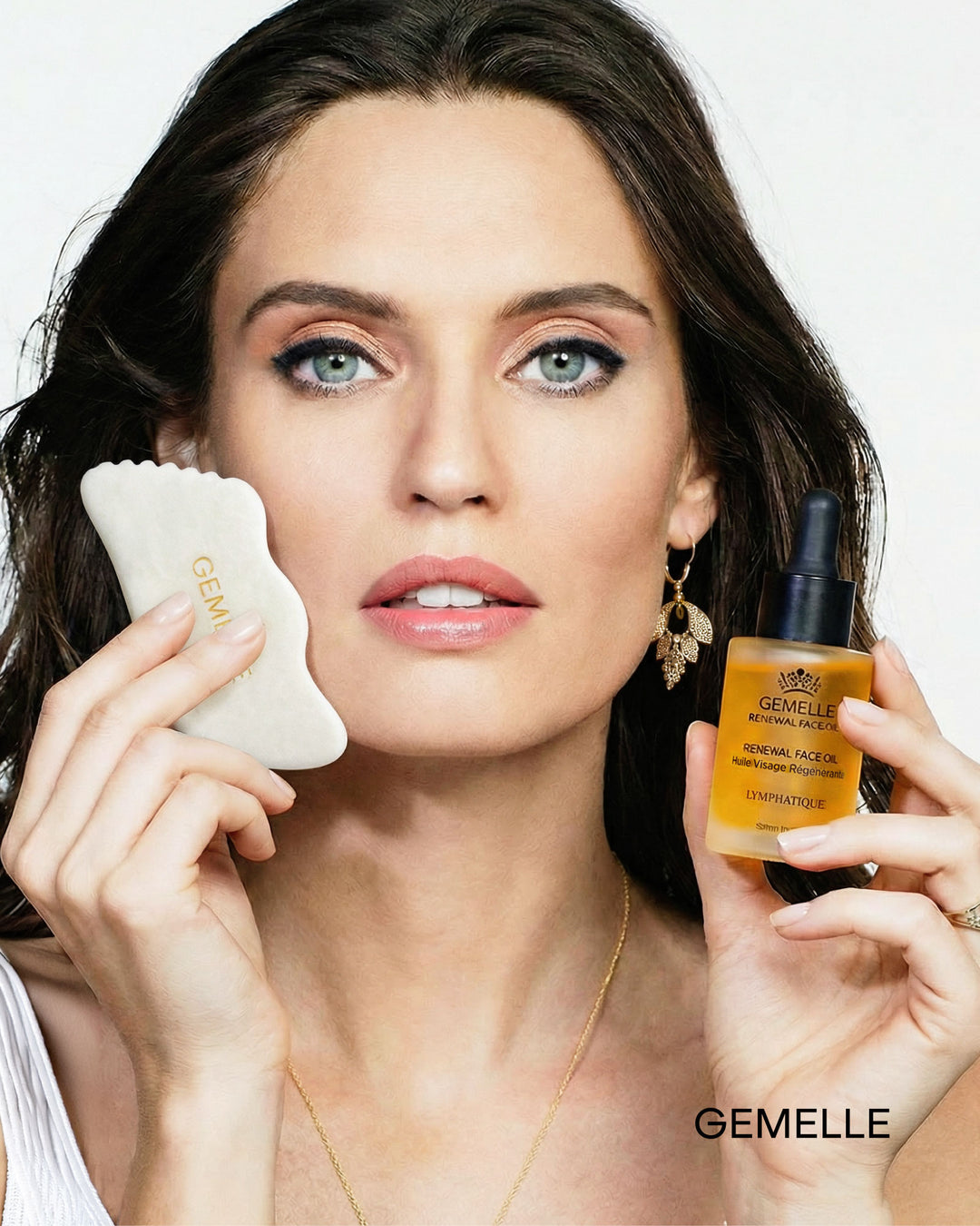 A GEMELLE Woman holding a White Jade Gua Sha facial tool and a bottle of GEMELLE Signature face oil skincare product against a white background